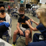 A masked melanated woman smiles with her eyes as a masked younger person looks on as they just received a COVID-19 vaccination at one of the City of Seattle vaccination sites. A blond-haired nurse is applying a band aid to this person's shoulder.