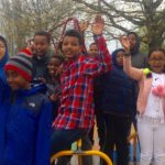 Image of a multi-racial group of smiling young people standing on a playground spinner with their hands in the air.