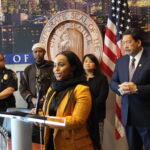 OIRA Director Mohamed speaking at podium at City Hall with Mayor Harrell and others standing behind her.
