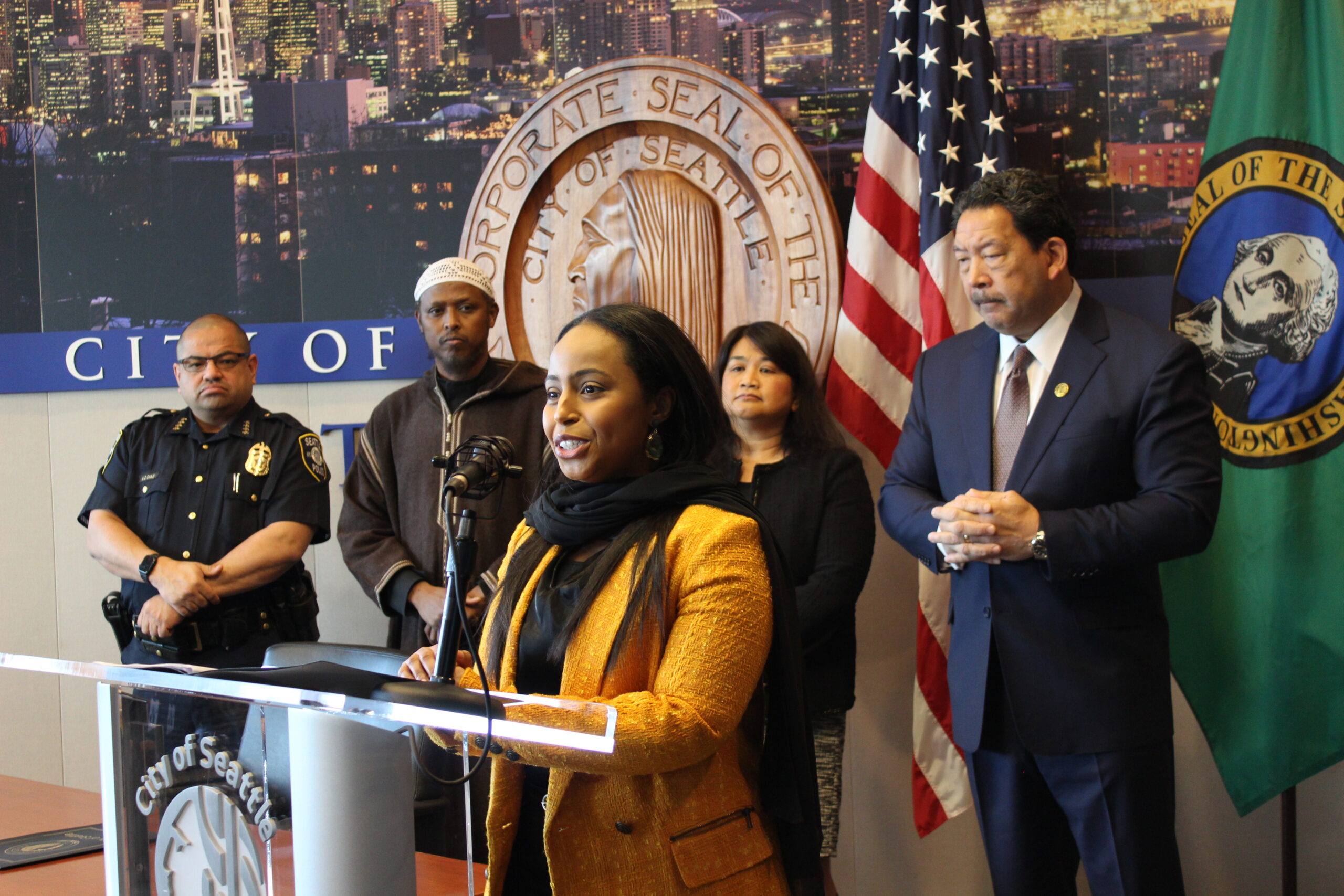 OIRA Director Mohamed speaking at podium at City Hall with Mayor Harrell and others standing behind her.