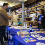 Woman standing behind a table with resources and flyers at a resource fair speaking to a man wearing a backpack.