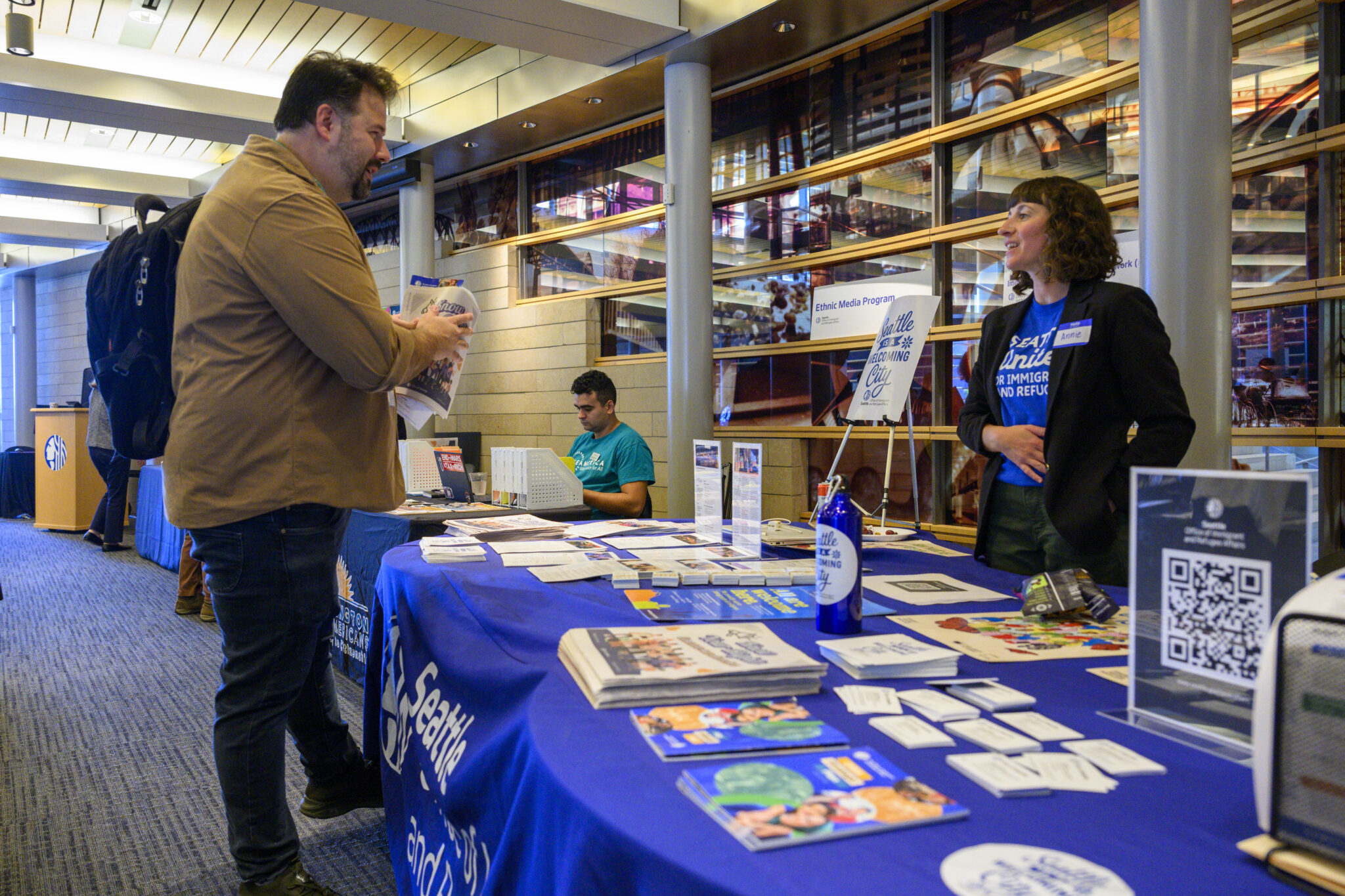 Woman standing behind a table with resources and flyers at a resource fair speaking to a man wearing a backpack.